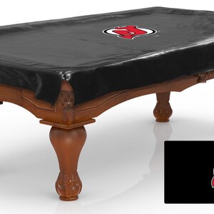 May include: Black pool table cover with the New Jersey Devils logo in red. The cover is made of a durable material and is designed to protect your pool table from dust and dirt.