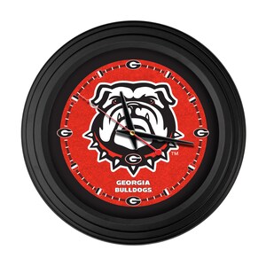 May include: Round black wall clock featuring the Georgia Bulldogs logo in the center. The clock face is red with white and black accents, including the team name and logo. The clock hands are black and red.