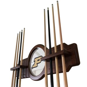 May include: A wooden pool cue rack with a circular mirror featuring a gold and black "P" logo. The rack holds multiple pool cues, some black and some light brown. The rack is made of dark brown wood.