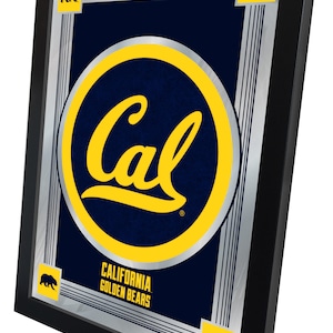 May include: A framed mirror with a silver background and a blue and yellow logo that says "Cal" in a circle. The logo is surrounded by a silver border. The text "California Golden Bears" is below the logo. There are four small bear silhouettes in the corners of the frame.