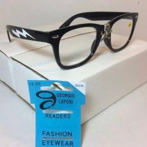 May include: Black plastic reading glasses with a skull and crossbones design on the side. The glasses have a white lightning bolt design on the arms. The glasses are on a white background with a blue and white price tag that reads "Giorgio Caponi Readers Fashion Eyewear +1.50 $14.99".