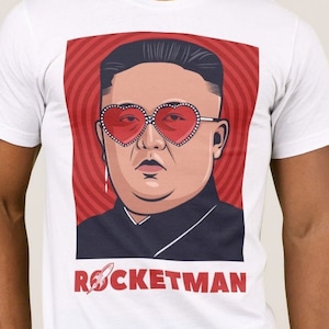 May include: A white t-shirt with a red, white, and blue graphic of a man wearing heart-shaped sunglasses. The graphic has a red background with a white circle pattern. The text "ROCKETMAN" is below the graphic with a rocket ship icon.