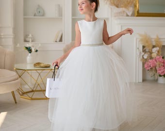 May include: A young person wearing a white sleeveless dress with a tulle skirt and a beaded belt. The dress is paired with matching shoes. The person is holding a white shopping bag with black handles and the word "L.NOV" printed on it.