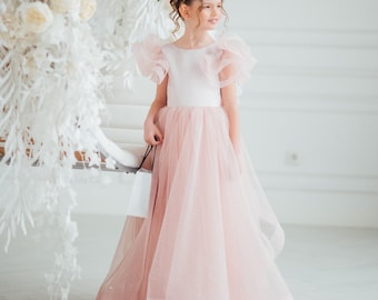 May include: A young girl wearing a light pink tulle dress with a white bodice and ruffled sleeves. The dress has a full skirt and is floor-length. The girl is smiling and holding a small white bag. The background is a bright white room with floral decorations.
