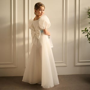 May include: A white formal dress with a large bow on the back and a light blue sash. The dress features puffy sleeves and a flowing skirt. The girl is looking over her shoulder.