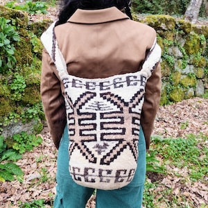 Puede incluir: Bolso de hombro tejido con un patrón geométrico en crema, marrón y negro. El bolso se lleva sobre el hombro, con una chaqueta marrón y pantalones verde azulado visibles. El fondo muestra un muro de piedra y follaje.