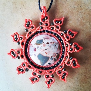 Puede incluir: Un colgante de sol de macramé rojo y negro con una piedra redonda moteada en el centro.