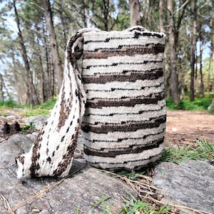 May include: A cylindrical woven bucket bag featuring a cream, dark brown, and black horizontal striped pattern. The bag includes a wide, matching woven shoulder strap with a similar textured design, crafted from thick, durable fiber material.
