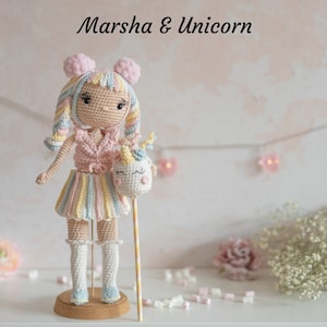 May include: A crocheted doll with pink and yellow striped skirt and a unicorn head on a stick. The doll has pink hair and a pink and white crocheted top. The doll is standing on a wooden base. The text "Marsha & Unicorn" is written above the doll.