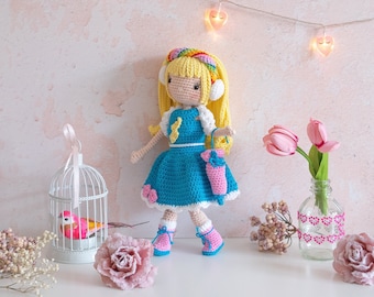 Pode incluir: Uma boneca de crochê com cabelo loiro vestindo um vestido azul e segurando um guarda-chuva rosa. A boneca está em pé sobre uma superfície branca com uma gaiola de pássaros e flores.