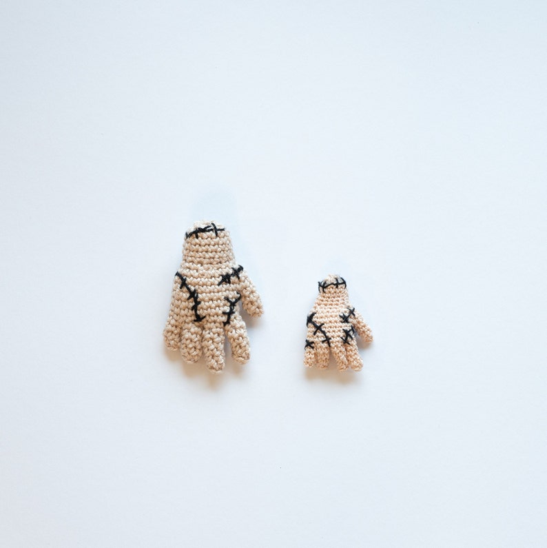 May include: Two crocheted hands with black stitching, one larger than the other, on a white background.