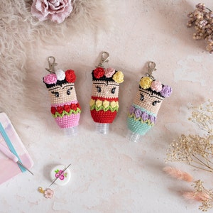 Könnte beinhalten: Drei gehäkelte Handdesinfektionsflaschen-Anhänger mit Schlüsselringen. Jeder Anhänger hat eine bunte Blumenkrone und ein lächelndes Gesicht. Die Anhänger sind rosa, rot und grün.
