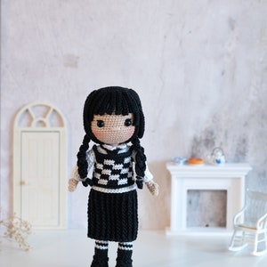 May include: A crocheted doll with black hair, a white and black checkered vest, a black skirt, and black boots. The doll is standing on a white surface.