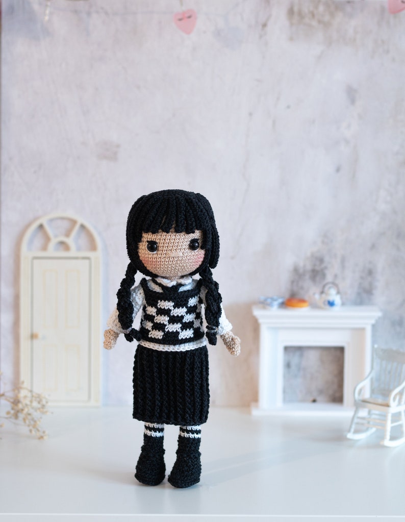 May include: A crocheted doll with black hair, a white and black checkered vest, a black skirt, and black boots. The doll is standing on a white surface.