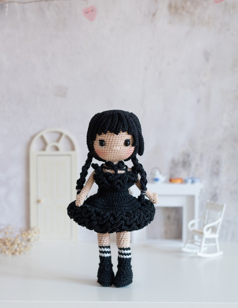 May include: A black crocheted doll with long black hair, wearing a black dress with a ruffled hem and black and white striped socks. The doll is standing on a white surface.