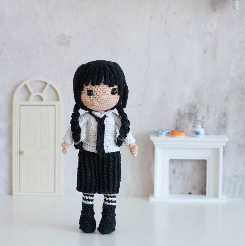 May include: A crocheted doll wearing a white shirt with a black tie, a black skirt with white stripes, and black and white striped socks. The doll has black hair and a serious expression.