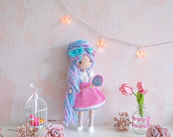 Pode incluir: Uma boneca de crochê com cabelo longo azul e rosa, vestindo um vestido rosa e segurando uma pirulito. A boneca está em pé sobre uma superfície branca com um fundo rosa e luzes de corda com lâmpadas em forma de coração.