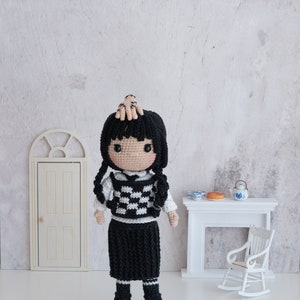 May include: A crocheted doll with black hair, a white and black checkered vest, a black skirt, and black boots. The doll is standing on a white surface in front of a white fireplace with a rocking chair.