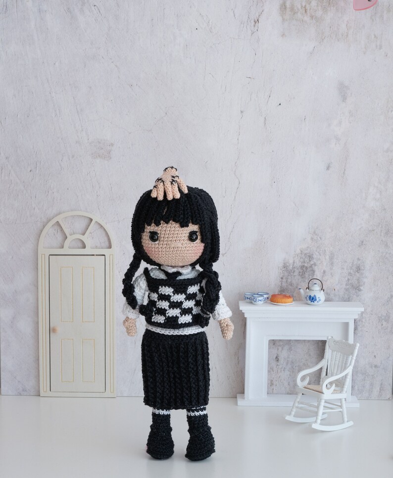 May include: A crocheted doll with black hair, a white and black checkered vest, a black skirt, and black boots. The doll is standing on a white surface in front of a white fireplace with a rocking chair.