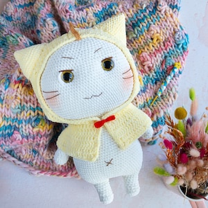 May include: A handmade white crochet cat doll wearing a yellow hooded cape and a red bow tie. The doll has embroidered facial features and is holding a small wand. The background includes a colourful knitted blanket and a bouquet of dried flowers.