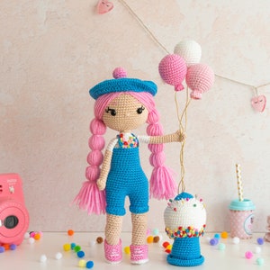 May include: A crocheted doll with pink hair and a blue beret holds a bunch of pink and white balloons. The doll is wearing a blue overall and is standing next to a crocheted gumball machine.