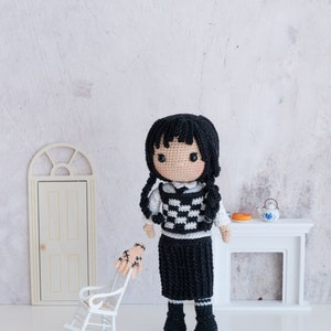 May include: A crocheted doll with black hair, a white and black checkered vest, and a black skirt. The doll is wearing black shoes and is standing in front of a white fireplace.