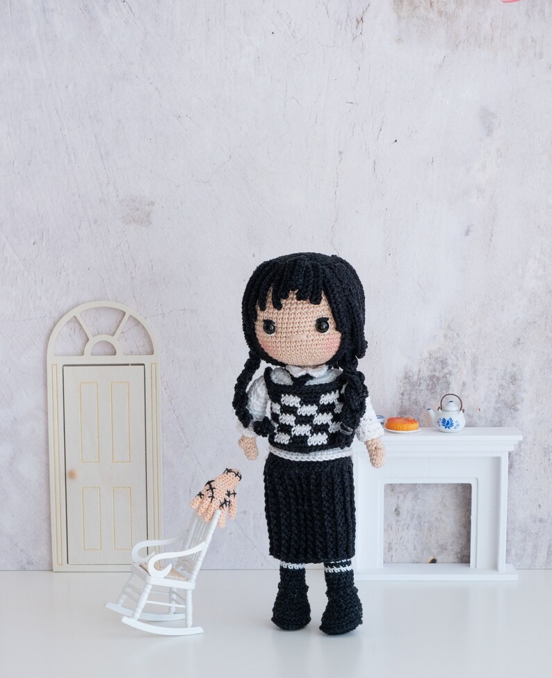 May include: A crocheted doll with black hair, a white and black checkered vest, and a black skirt. The doll is wearing black shoes and is standing in front of a white fireplace.