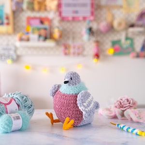 May include: A handmade, plush pigeon in shades of gray, pink, and teal. The bird has orange feet and a black beak. A ball of light blue yarn and a crochet hook are next to the pigeon. The background is a blurred display of craft supplies.