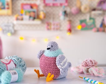 以下が含まれることがあります： グレー、ピンク、ティール色の手作りのぬいぐるみハト。鳥はオレンジ色の足と黒い嘴を持っています。水色の毛糸玉と編み棒がハトの隣にあります。背景は、ぼやけたクラフト用品のディスプレイです。