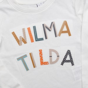 Könnte beinhalten: Weißes Langarmshirt mit den Namen "WILMA" und "TILDA", die in bunten, gemusterten Buchstaben geschrieben sind.