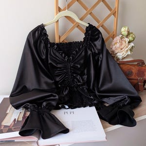 Könnte beinhalten: Schwarze Satinbluse mit langen, weiten Ärmeln und gerafftem Oberteil. Die Bluse hängt an einem weißen Kleiderbügel. Das Wort "PUPE" ist auf einem Buch zu sehen. Im Hintergrund ist ein Holzgitter zu sehen.