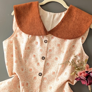 Puede incluir: Una blusa sin mangas de tela rosa claro con un patrón de rosas repetitivo, acentuada por un cuello grande y ancho de pana color óxido. La parte delantera de la prenda se cierra con una fila vertical de tres botones circulares oscuros.