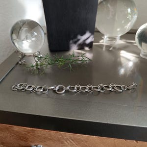 Könnte beinhalten: Ein silberfarbenes Armband mit einer Reihe von miteinander verbundenen ovalen und kreisförmigen Gliedern. Das Armband hat einen Verschluss und wird auf einer grauen Oberfläche präsentiert. Der Schmuck ist ein schlichtes, elegantes Accessoire.