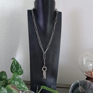 Könnte beinhalten: Eine silberfarbene Halskette mit einem Schlüsselanhänger, präsentiert auf einem schwarzen Schmuckständer. Die Kette hat längliche Glieder, und der Schlüsselanhänger hängt am Ende. Im Hintergrund sind eine Pflanze und eine Glaskugel zu sehen.