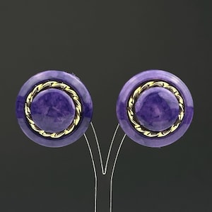 Peut inclure: Une paire de boucles d'oreilles rondes à clip en violet et or. Les boucles d'oreilles ont un centre violet texturé avec une bordure en or en forme de corde.