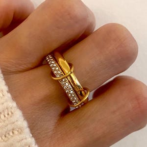 Könnte beinhalten: Ein goldener Ring mit einem Doppelband-Design. Ein Band ist aus einfachem Gold, das andere ist mit kleinen, klaren Steinen besetzt.