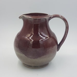 Może przedstawiać: Ceramiczny dzbanek z zaokrąglonym korpusem i uchwytem. Dzbanek jest pokryty glazurą w kolorze ciemnego bordo z jaśniejszą brązową glazurą na dole.