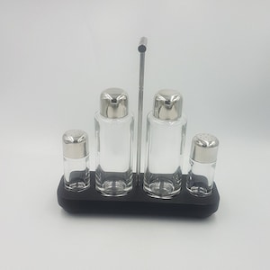 May include: A set of four glass condiment bottles with silver lids and a black metal stand. The bottles are arranged in a row on the stand, with the two larger bottles in the centre and the two smaller bottles on either side.