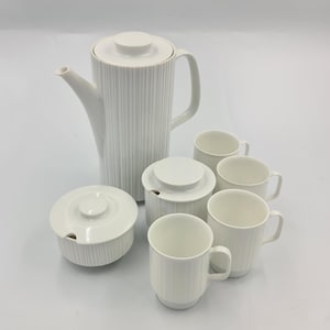 Könnte beinhalten: Ein weißes Keramik-Kaffeeservice mit geripptem Design. Das Set umfasst eine Kaffeekanne, eine Zuckerdose, einen Sahnekännchen und vier Tassen. Die Kaffeekanne hat einen Deckel und einen Griff. Die Tassen haben Griffe. Das Set steht auf einer weißen Oberfläche.