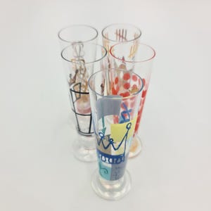 Puede incluir: Cinco copas transparentes con diseños coloridos pintados a mano. Las copas presentan varios patrones, incluyendo puntos, líneas y formas geométricas en rojo, azul, amarillo y negro. Las copas están dispuestas sobre una superficie blanca.