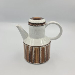 May include: A ceramic teapot with a white speckled body, a brown and white striped base, and a matching lid. The teapot has a curved handle and a spout for pouring. The design is a mix of neutral tones and vertical stripes.