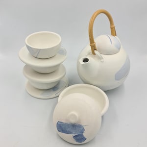 Könnte beinhalten: Ein weißes Keramik-Teeservice mit blauen Pinselstrich-Akzenten. Das Set beinhaltet eine Teekanne mit Bambusgriff, einen Stapel kleiner Untertassen und Schalen sowie einen Teekannendeckel.