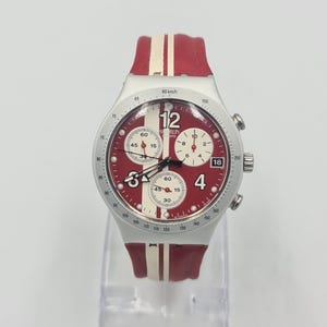 May include: A red and white Swatch chronograph watch with a silver case. The watch features a red dial with white accents and a red and white striped silicone band. The watch has a 12, 3, 4, and 18 hour marker.