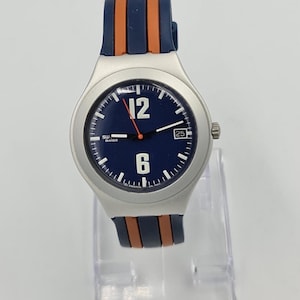 May include: Silver-toned wristwatch with a navy blue dial featuring white numerals at 12 and 6 o'clock. The watch has a date display at the 3 o'clock position and a navy blue and orange strap. The watch is displayed on a clear stand.