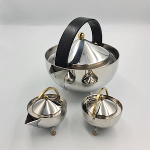 May include: A stainless steel tea set with a teapot and two smaller serving pieces. The teapot has a black handle and a gold-colored finial. The serving pieces have gold-colored handles and small gold-colored feet. The set is on a white background.