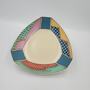 May include: A cream-coloured, triangular plate with a colourful geometric border. The border features teal, pink, yellow, and orange sections with checkered and striped patterns. The plate is designed with a modern, artistic aesthetic.