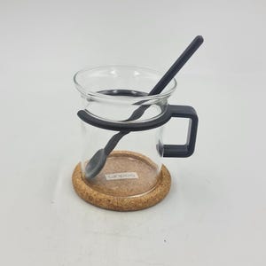 May include: A clear glass mug with a black handle and a black spoon inside. The mug sits on a round cork coaster. The mug has a black band around the top and bottom. The brand name "bodum" is printed on the coaster.
