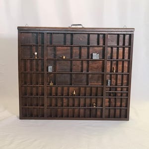 May include: A rectangular dark brown wooden printer's tray featuring numerous small, varied compartments arranged in a grid. Several tiny metal objects, including a small pitcher, a letter block with the letters WO, and other trinkets, are placed inside.