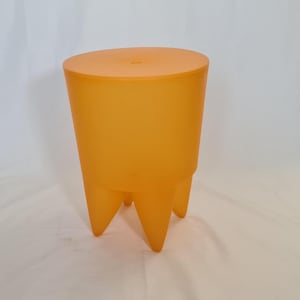 Könnte beinhalten: Ein durchscheinender orangefarbener Hocker mit zylindrischem Körper und flacher, runder Oberseite. Der Hocker hat drei konische Beine. Das Design ist modern und minimalistisch, geeignet für den Innen- oder Außenbereich.