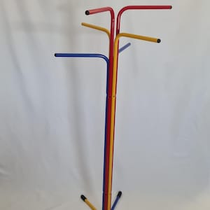 May include: A freestanding coat rack constructed from tubular metal in primary colors. The structure features a central vertical pole with multiple extending arms at the top for hanging items and a four-legged base, all finished with black end caps.
